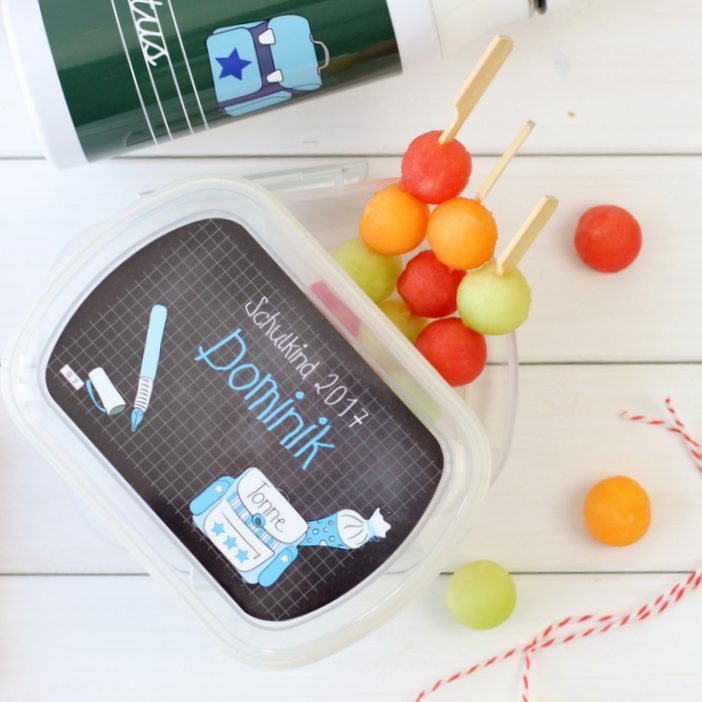 Eine Brotdose mit bunten Melonenkugeln auf Spießen. Motiv auf der Dose ist ein blauer Schulranzen, eine Schultüte und ein Stift.