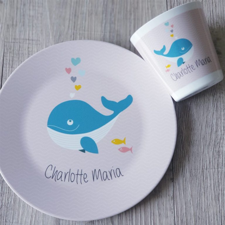 Ein Teller und ein Becher mit einem blauen Wal auf rosa Hintergrund und dem Namen "Charlotte Mana".